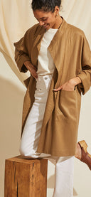 Femme en veste longue camel et pantalon blanc, un pied posé sur un tabouret en bois