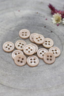Douzaine de petits boutons ronds à quatre trous couleur rose blush pâle brillant, empilés sur pierre grise avec fleurs séchées en haut à droite.
