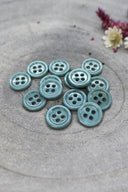 Douzaine de petits boutons ronds à quatre trous couleur vert cactus opaque brillant, empilés sur pierre grise avec fleurs séchées en haut à droite.