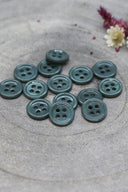 A dozen small four-hole round buttons in glossy greyish cedar green, stacked on grey stone with dried flowers.