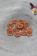 Douzaine de petits boutons ronds à quatre trous couleur châtaigne (marron orangé) brillant, empilés sur fond de pierre grise avec fleurs séchées en arrière-plan.
