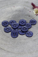 A dozen small four-hole round buttons in deep glossy cobalt blue, stacked on grey stone with dried flowers in the background.