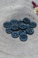 A dozen small four-hole round buttons in glossy bluish forest green, stacked on grey stone with dried flowers.