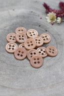 A dozen small four-hole round buttons in glossy sandy beige-pink, stacked on grey stone with dried flowers in the upper right.