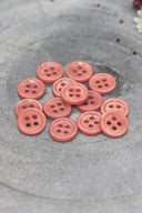 A dozen small four-hole round buttons in glossy peachy pink (melba), stacked on grey stone with dried flowers in the upper right.