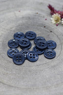 Douzaine de petits boutons ronds à quatre trous bleu marine foncé (bleu nuit) brillant, empilés sur pierre grise avec fleurs séchées.