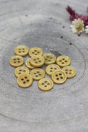 A dozen small four-hole round buttons in glossy mustard yellow, stacked on grey stone with white and pink dried flowers in the upper right.