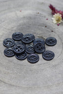 Douzaine de petits boutons ronds à quatre trous gris anthracite très foncé, presque noir, empilés sur pierre grise avec fleurs séchées.