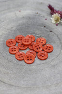 Douzaine de petits boutons ronds à quatre trous couleur orange vif (poppy tangerine) brillant, empilés sur pierre grise avec fleurs séchées en arrière-plan.