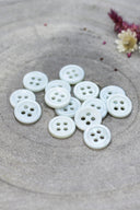 A dozen small four-hole round buttons in pearly white with a subtle pale green sheen, stacked on grey stone with dried flowers.