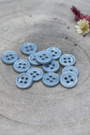 A dozen small four-hole round buttons in glossy light blue-grey (storm), stacked on grey stone with dried flowers.