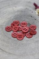 A dozen small four-hole round buttons in glossy terracotta (brick red), stacked on grey stone with dried flowers.