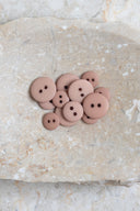 Boutons ronds classiques à deux trous, finition mate rose taupe blush, présentés en tas sur une pierre beige marbrée.