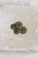 Boutons ronds classiques à deux trous, finition mate vert olive kaki, présentés en tas sur une pierre beige marbrée.