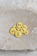 Boutons ronds classiques à deux trous, finition mate jaune citron pâle, présentés en tas sur une pierre beige marbrée.