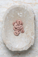Boutons ronds classiques à deux trous, finition mate beige rosé sable, présentés en tas sur une pierre beige marbrée.