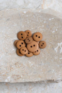 Boutons ronds classiques à deux trous, finition mate ocre caramel, présentés en tas sur une pierre beige marbrée.