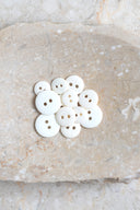 Boutons ronds classiques à deux trous, finition mate blanc cassé, présentés en tas sur une pierre beige marbrée.
