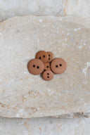 Boutons ronds classiques à deux trous, finition mate brun caramel pecan pie, présentés en tas sur une pierre beige marbrée.