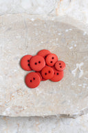 Boutons ronds classiques à deux trous, finition mate rouge orangé tangerine, présentés en tas sur une pierre beige marbrée.