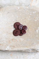 Boutons ronds classiques à deux trous, finition mate bordeaux brun rouille, présentés en tas sur une pierre beige marbrée.