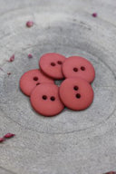 Boutons ronds classiques à deux trous, finition mate rouge terracotta, présentés en tas sur une pierre grise, avec une feuille séchée en arrière-plan.