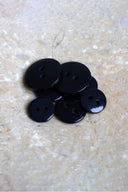 Boutons ronds classiques à deux trous, finition brillante noir profond, présentés en tas sur une pierre beige marbrée.