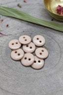 Boutons ronds classiques à deux trous, finition brillante rose taupe blush, présentés en tas sur une pierre grise, avec une feuille verte et des pétales séchés en arrière-plan.