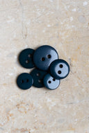 Boutons ronds classiques à deux trous, finition brillante bleu-vert forêt très foncé, présentés en tas sur une pierre beige marbrée.