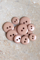 Boutons ronds classiques à deux trous, finition brillante beige rosé sable, présentés en tas sur une pierre beige marbrée.