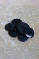 Boutons ronds classiques à deux trous, finition brillante noir intense (night), présentés en tas sur une pierre beige marbrée.