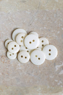 Boutons ronds classiques à deux trous, finition brillante blanc cassé nacré, présentés en tas sur une pierre beige marbrée.