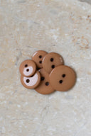 Boutons ronds classiques à deux trous, finition brillante brun caramel pecan pie, présentés en tas sur une pierre beige marbrée.