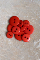 Boutons ronds classiques à deux trous, finition brillante rouge orangé tangerine, présentés en tas sur une pierre beige marbrée.