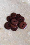 Boutons ronds classiques à deux trous, finition brillante bordeaux brun rouille, présentés en tas sur une pierre beige marbrée.