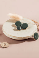 Domed round and oval buttons, glossy finish, in greyish cedar green, with one shown from the back revealing the metal shank fastening, on a white ceramic dish, pink background with a dried lagurus spike.