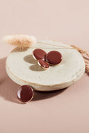 Quatre boutons ronds et ovales bombés, finition brillante, coloris marron chocolat foncé, cerclés d'un fin liseré doré, présentés sur une petite coupelle en céramique blanche, sur fond rose avec un épi de lagurus séché.
