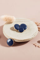 Quatre boutons ronds et ovales bombés, finition brillante, coloris bleu marine cobalt, cerclés d'un fin liseré doré, présentés sur une petite coupelle en céramique blanche, sur fond rose avec un épi de lagurus séché.