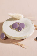 Four domed round and oval buttons, glossy finish, in pale mauve purple, rimmed with a thin gold trim, displayed in a small white ceramic dish on a pink background with a dried lagurus spike.
