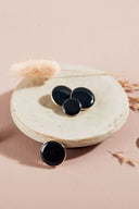 Quatre boutons ronds et ovales bombés, finition brillante, coloris noir profond à reflets verts (forêt), cerclés d'un fin liseré doré, présentés sur une petite coupelle en céramique blanche, sur fond rose avec un épi de lagurus séché.