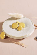 Four domed round and oval buttons, glossy finish, in lemon yellow, rimmed with a thin gold trim, displayed in a small white ceramic dish on a pink background with a dried lagurus spike.