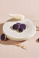 Quatre boutons ronds et ovales bombés, finition brillante, coloris violet profond, cerclés d'un fin liseré doré, présentés sur une petite coupelle en céramique blanche, sur fond rose avec un épi de lagurus séché.