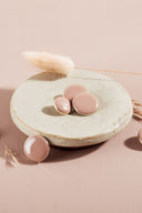 Quatre boutons ronds et ovales bombés, finition brillante, coloris beige rosé nude, cerclés d'un fin liseré doré, présentés sur une petite coupelle en céramique blanche, sur fond rose avec un épi de lagurus séché.