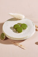 Quatre boutons ronds et ovales bombés, finition brillante, coloris vert matcha, cerclés d'un fin liseré doré, présentés sur une petite coupelle en céramique blanche, sur fond rose avec un épi de lagurus séché.