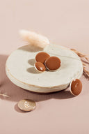 Boutons ronds et ovales bombés, finition brillante, coloris marron caramel, cerclés d'un fin liseré doré, dont un présenté de dos laissant voir l'attache à queue métallique, sur une coupelle en céramique blanche, fond rose avec un épi de lagurus séché.