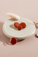 Quatre boutons ronds et ovales bombés, finition brillante, coloris orange rouge vif, cerclés d'un fin liseré doré, présentés sur une petite coupelle en céramique blanche, sur fond rose avec un épi de lagurus séché.