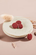 Quatre boutons ronds et ovales bombés, finition brillante, coloris rouge terracotta framboise, cerclés d'un fin liseré doré, présentés sur une petite coupelle en céramique blanche, sur fond rose avec un épi de lagurus séché.