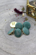 Five faceted round buttons in cactus green rimmed with a gold trim, with a metal shank, set on grey stone with dried flowers and a blurred gold box in the background.