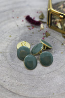 Five faceted round buttons in cedar green (dark sage green) rimmed with a gold trim, set on grey stone with dried flowers and a blurred gold box in the background.