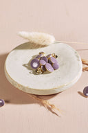 Domed round buttons in pale mauve purple rimmed with a thin gold trim, with a metal shank, piled in a small white ceramic dish on a pink background, with a sprig of dried lagurus grass in the background.